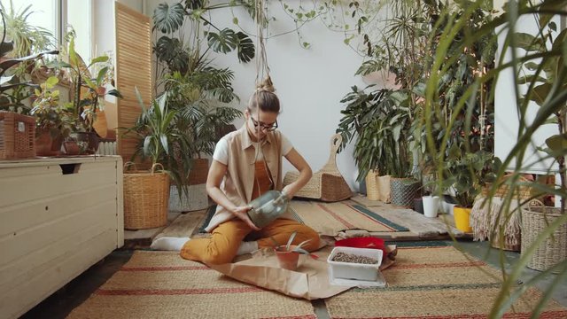 Young woman with hair bun sitting on the floor in her indoor garden, taking soil from glass jar and filling container with it while planting flower in pot