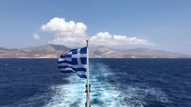A Greek flag flapping with Serifos island, blue sea and sky, white clouds and foam in background.