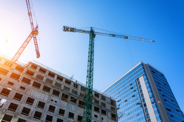 Bottom view of a building under construction with high construction cranes and a glass tower in the background.