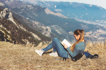 Naklejka na ściany i meble Young beautiful girl travels alone in the mountains in spring or autumn, sits on the edge of the mountain and reading book and enjoys nature, rocks and green forests, view of the landscape. a backpack