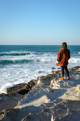 Person standing on the edge of rocky cliff, looking at the waves in the ocean. View from the back.