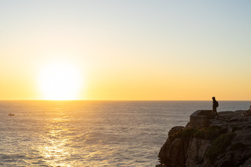 A person in the distance standing on the edge of a cliff looking at the ocean in the sunset. View from the side.