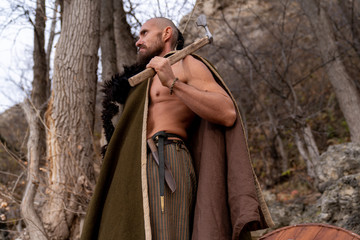 Viking dressed in a cape and skin armed with an ax and a sword stands on the background of the forest