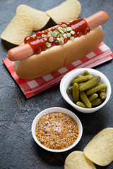 Close-up of a hot-dog with potato chips, pickles and mustard on a dark brown stone surface,...