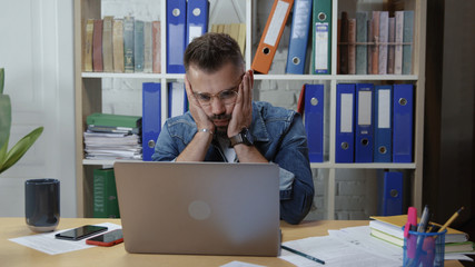 Unmotivated young man sitting at desk with laptop feeling strees and disinterest in work. Boring profession.