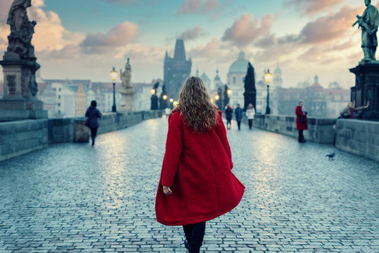 Woman in red coat walking on The Charles Bridge in Prague during the atmospheric sunset in winter