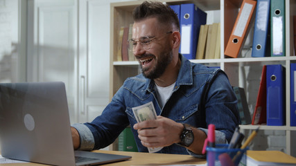 Excited young successful businessman making money online getting banknotes from laptop in surprise....