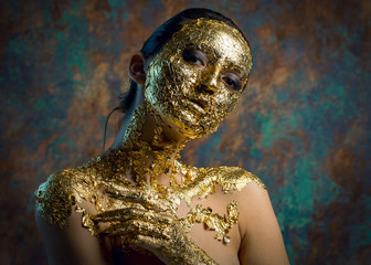 Girl with a mask on her face made of gold leaf. Gloomy studio portrait of a brunette on an abstract background.