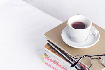 Stack of different books and notepads with woman's glasses and coffee cup on the top on a white table.