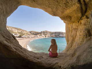 Beautiful hippie woman siting in the cave by the sandy beach. Holiday in Matala on Crete, Greece.