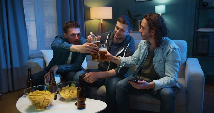 Three Caucasian guys taking their glasses with beer and cheers with it when sitting on sofa late in evening at home. Young men watching TV together at night with snacks.