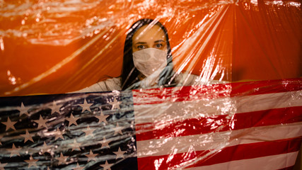 Young woman in medical mask with American flag in isolation from coronovirus on orange background. Portrait of female covering her face with mask to protect yourself from diseases. Concept of epidemic