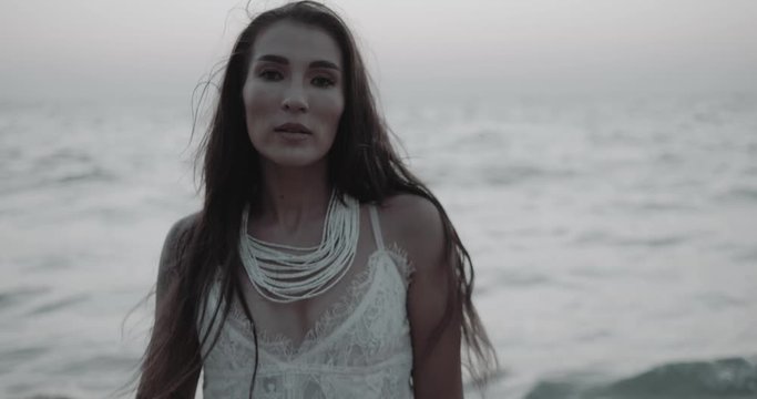 Romantic portrait of beautiful brunette woman in white lace dress walking, flirting with the camera at the beach during summer early evening - video in slow motion