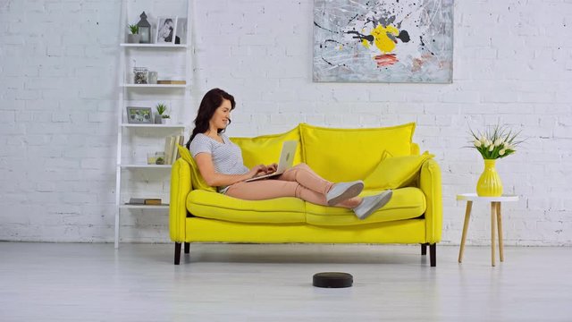 woman remote working with laptop near robotic vacuum cleaner