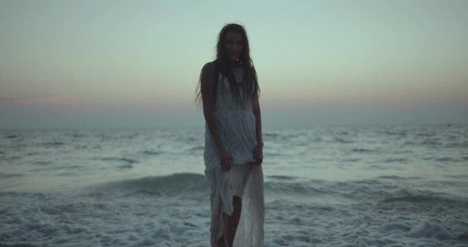 Romantic portrait of beautiful brunette woman in white lace dress walking, flirting with the camera at the beach during summer early evening - video in slow motion