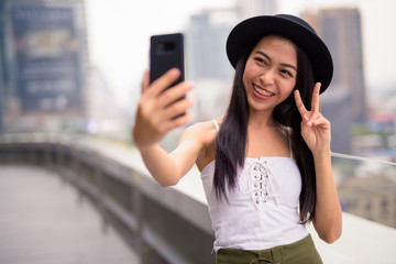 Happy young beautiful Asian tourist woman taking selfie against view of the city