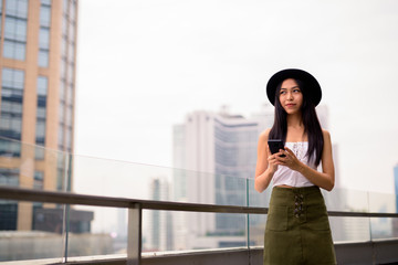 Young beautiful Asian tourist woman thinking while using phone against view of the city