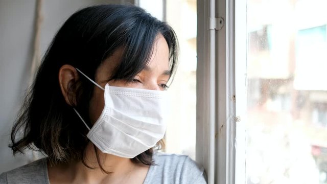 close up of young woman infected with corona virus with face mask looking out of window while in quarantine with sad and worried expression due to self isolation