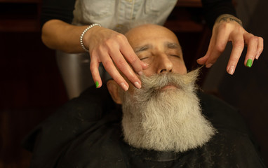 Handsome senior man getting styling and trimming of his beard in the barbershop.