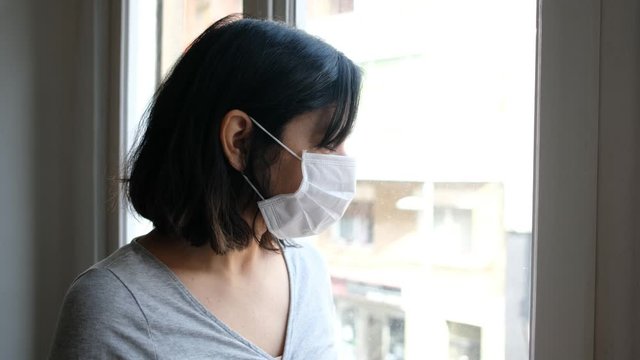 portrait of young woman infected with corona virus coughing with face mask looking out of window while in quarantine with sad and worried expression due to self isolation