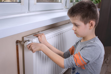 Chłopiec w wieku szkolnym trzyma pokrętło regulatora temperatury w kaloryferze i ustawia odpowiednią temperaturę. 