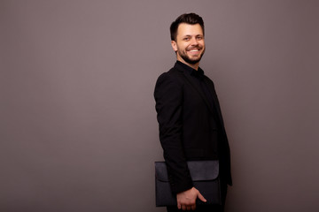 Obraz na płótnie Canvas Studio shoot. Portrait of smiling happy bearded businessman in black classic suit standing against grey wall. Young man holding black portfolio. Copy space for advertisement.