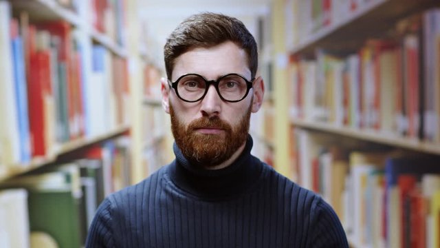 Portrait of intelligent caucasian young man wearing glasses looking smart confident posing in the raw of bookshelves at library. People on background. Education concept.
