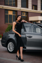 Fototapeta na wymiar Stylish young girl stands near the car in a black dress. Business fashion and style