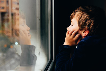 Sad and melancholic boy looks through a window in his apartment during his confinement