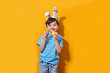 Portrait of a smiling adorable little boy with bunny ears and holding colorful easter eggs isolated orange background