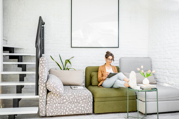 Young creative and well-dressed woman working on a digital tablet while sitting on the comfortable couch at home. Interior view on the modern and bright living room