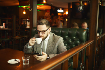Young businessman in eyeglasses and in suit drinking coffee and using his mobile phone while sitting at the table in cafe