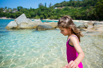 Cute happy little girl running along the beach in swimming suit, jumping over waves. Beautiful summer sunny day, clear turquoise sea, rocks, white sand, picturesque tropic landscape. Phuket, Thailand