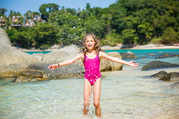 Cute happy little girl running, dancing on the beach in pink swimming suit. Beautiful summer sunny day, turquoise sea, rocks, white sand, picturesque tropic landscape. Phuket, Thailand