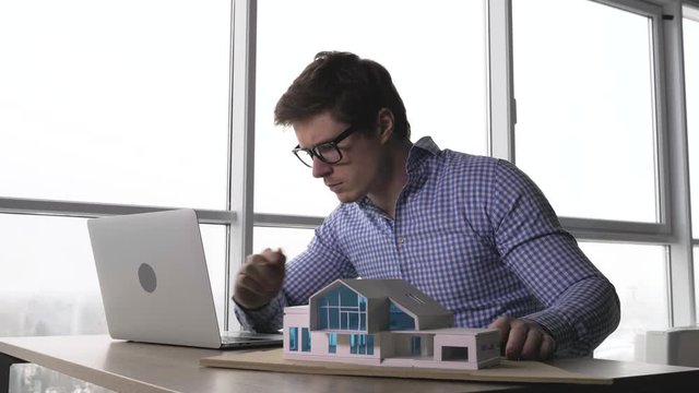 caucasian male architect working in the office in front of the laptop and examing house model