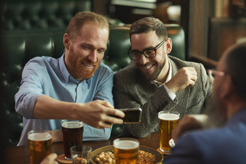 Two young bearded men making a selfie portrait on mobile phone while drinking beer at the table in the pub
