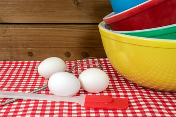 stack of retro mixing bowls on checkered tablecloth with white eggs and kitchen utensils