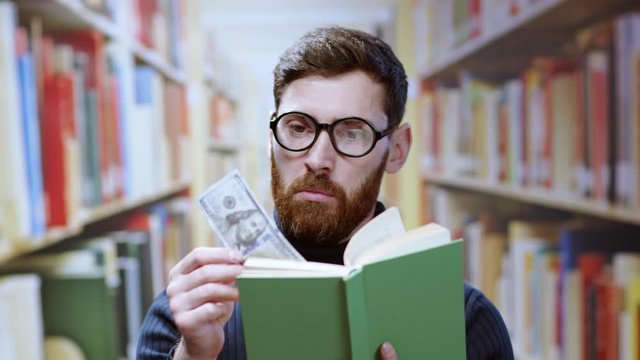 Lucky bearded hipster student finds a one hundred dollar banknote secret bookmark in a book staying surprised and excited in library or bookstore.
