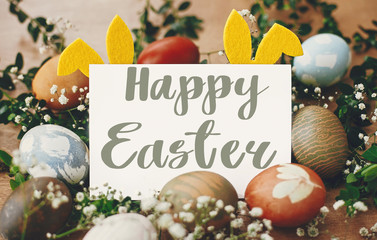 Happy Easter text. Easter greetings card. Stylish easter eggs, yellow bunny ears in nest of spring flowers, and card on rustic wooden table. Natural dyed easter eggs. Holiday card