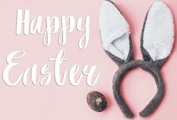 Happy Easter text. Easter greetings card. Stylish easter egg and bunny ears flat lay on pink wallpaper. Natural dyed marble easter egg and grey rabbit ears. Seasons greeting. Holiday card