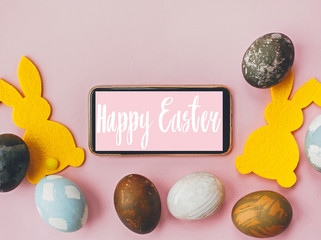 Happy Easter text. Easter greetings card. Phone with greeting, stylish easter eggs, yellow bunnies on pink paper flat lay. Modern natural dyed blue and marble easter eggs. Holiday card