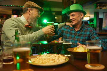 Two smiling friends talking and toasting with beer while sitting at the table with snacks in the bar