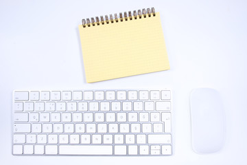 Top view of workplace desktop, there is a white keyboard, white mouse and yellow notebook or notepad