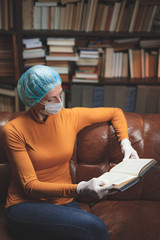 Obraz premium Woman with protective antiviral mask sitting at home in isolation / quarantine and reading book.
