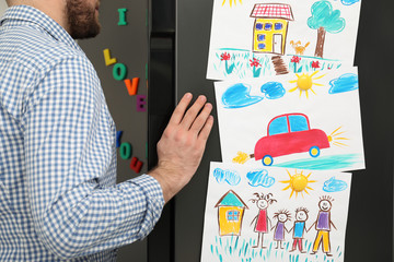 Man opening refrigerator door with child's drawings and magnets, closeup