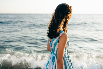 Rear view image of brunette beautiful woman walking along beach and sea at the sunset sunlight background. Pretty female wearing dress, walking outside on the ocean.