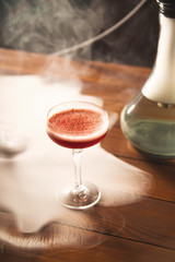 Raspberry alcoholic cocktail in a glass with foam sprinkled with freeze-dried raspberries in the hookah smoke on the table.