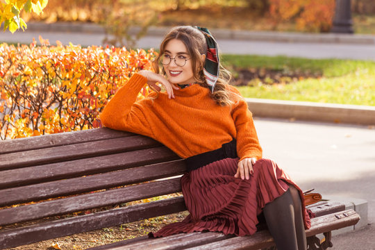 A beautiful, young woman sits on a bench in the fall, in October. The girl is dressed round, with glass glasses. Student teacher is resting, walking in the park.