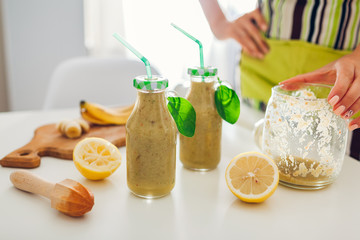 Bottles with spinach and banana smoothie with ingredients. Woman filled in and decorated drinks on kitchen table. Healthy detox diet