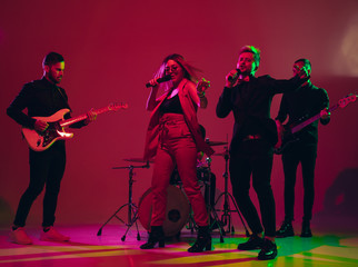 Young and joyful caucasian musicians performing on red studio background in neon light. Concept of music, hobby, festival. Colorful portrait of modern artist. Attented and inspired. Art, cover band.